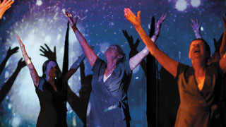 Several people standing with their arms raised, in front of a starry blue background. Light is projected onto them so their shadows appear on the background.