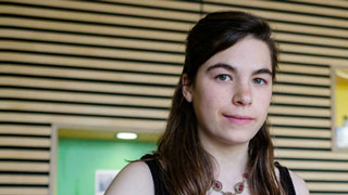 portrait of a young woman looking at the camera. She stands in front of an interior wall made of wooden slats.